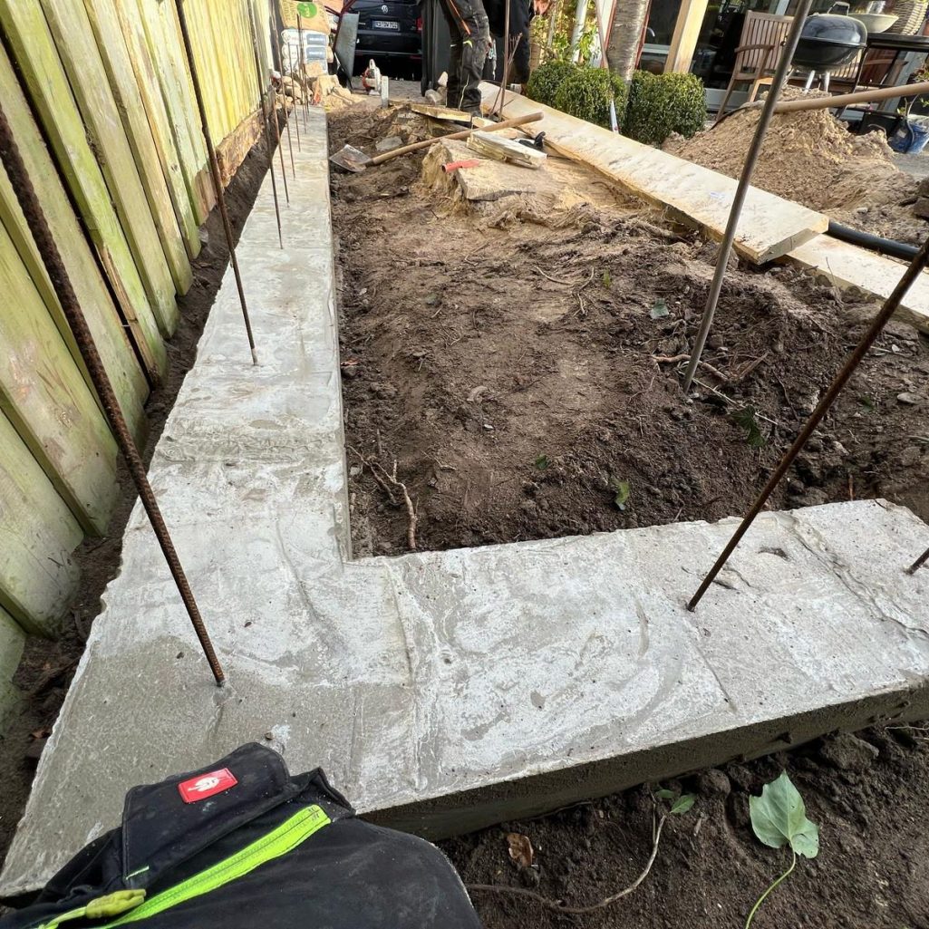 Ein Baustellenbereich mit einem L-förmigen, gegossenen Betonsockel und Bewehrungsstäben.